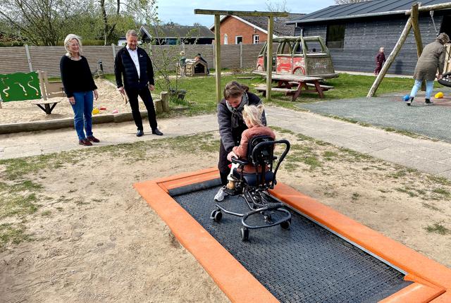 Pædagog Sofie Mølgaard Nieslen hjælper her et barn i kørestol ind på trampolinen. I baggrunden ses institutionens leder, Susanne Østergaard, og afdelingsdirektør i Sparekassen Danmark, Villy Sørensen.