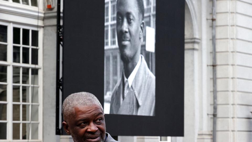 Roland Lumumba passerer en plakat med et foto af hans afdøde far, DR Congos uafhængighedshelt Patrice Lumumba, ved ankomsten til en ceremoni i Bruxelles mandag. Her udleverede Belgien en tand fra Lumumba til hans familie. <i>Pool/Reuters</i>