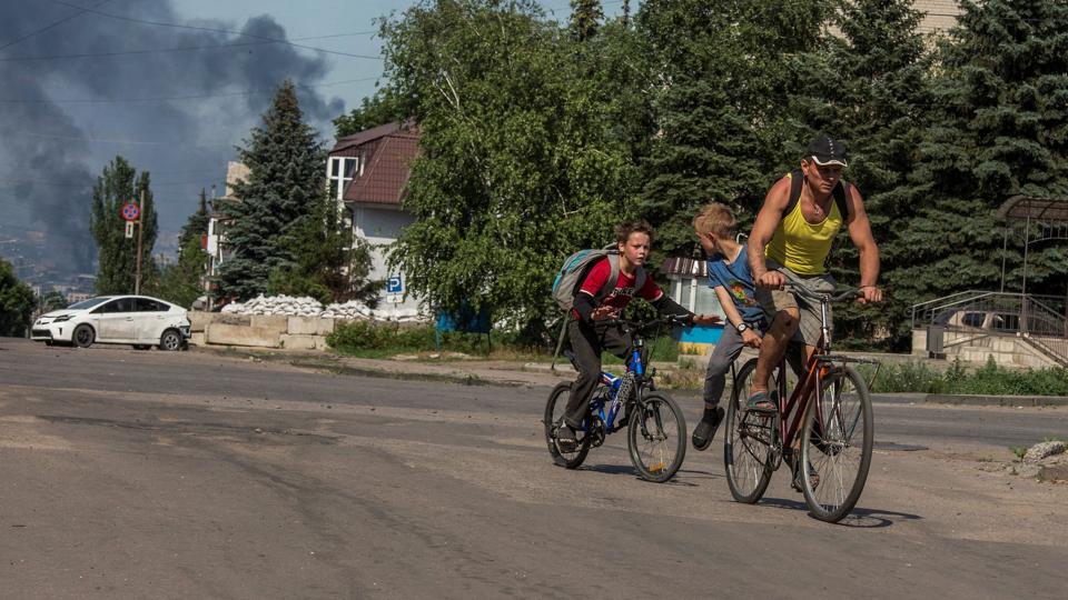 To børn og en mand cykler, mens røg fra et bombardement stiger op i baggrunden i den ukrainske by Lysytjansk i regionen Luhansk i juni i år. <i>Stringer/Reuters</i>