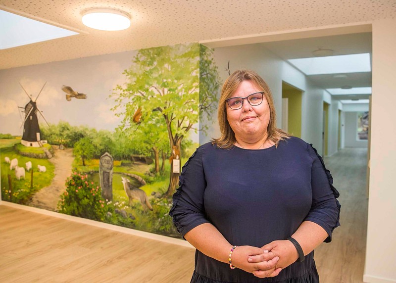 Plejehjemsleder Karin Nielsen glæder sig til at tage imod beboere, pårørende og kommende personale. I baggrunden et af de malerier som kunstner Kiss Jönsson har kreeret til stedet..