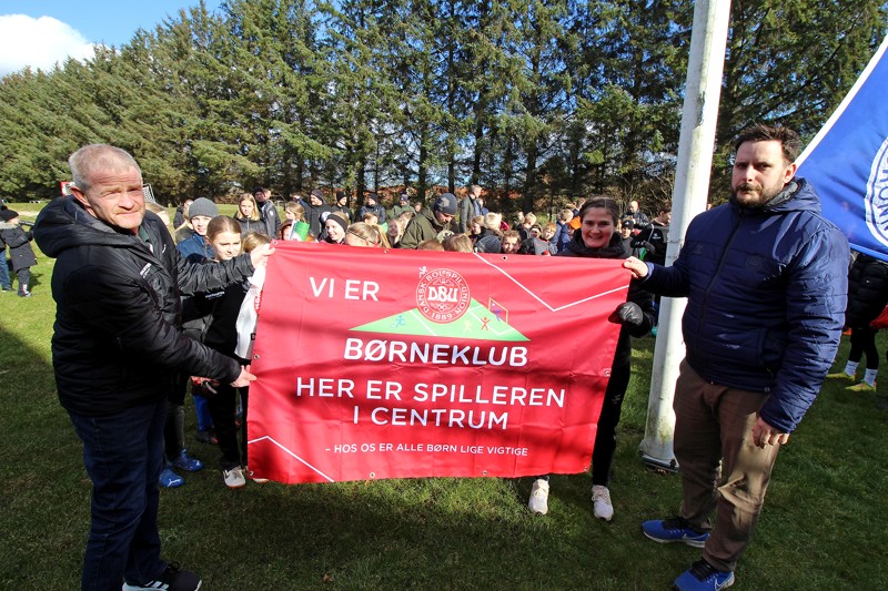 Det synlige bevis på, at klubben nu er DBU Børneklub. Til venstre ses klubbens næstformand, Anders Røge, og til højre Kresten Mathiassen fra DBU.